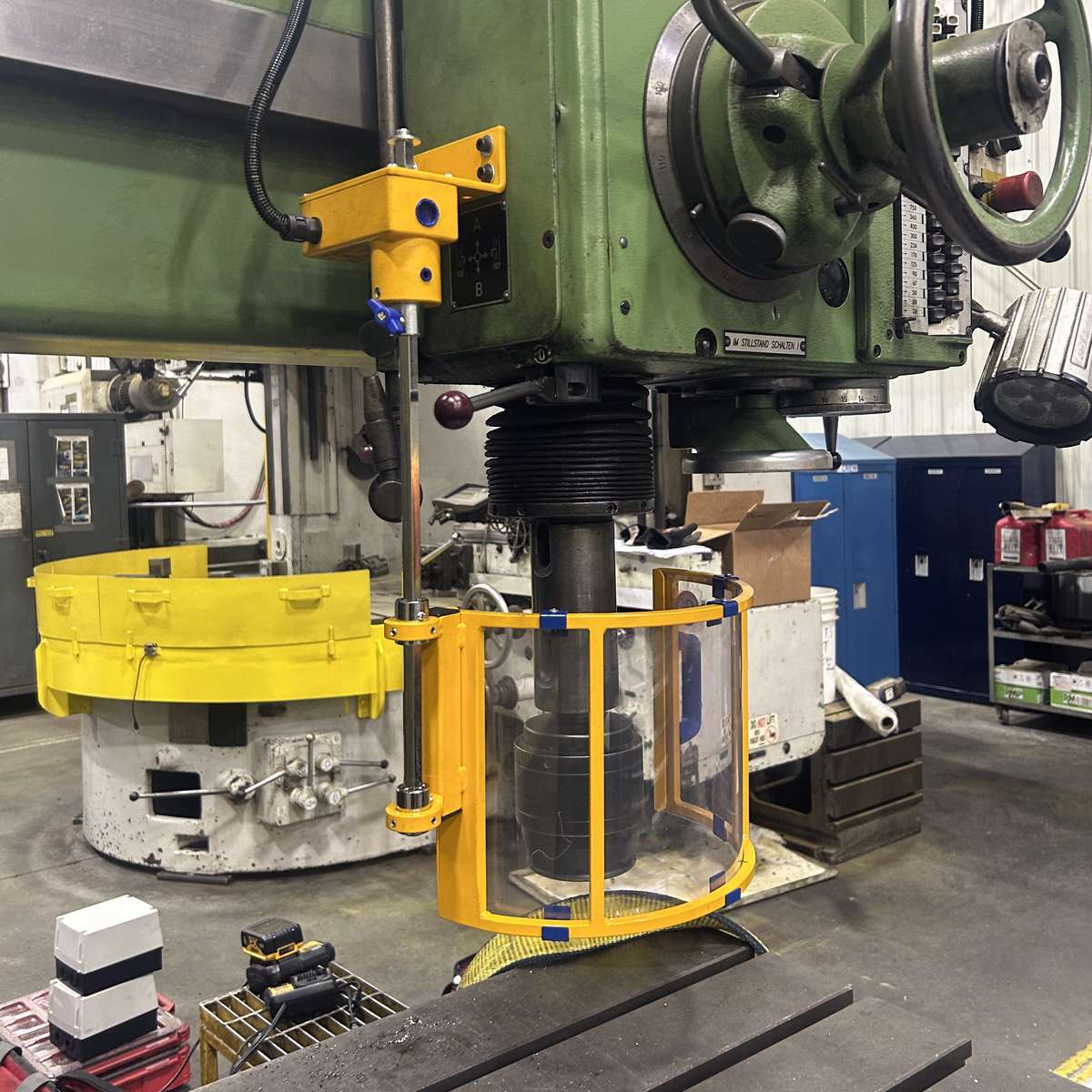 A safety guard installed on a radial drill press. The shield is mounted on the left side of the drill head.